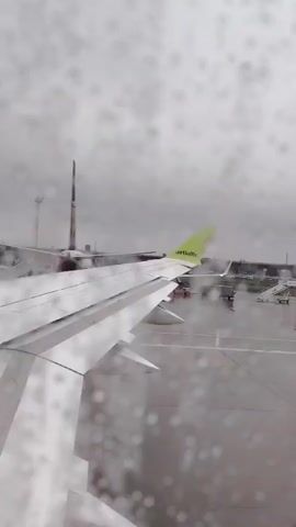 A vertical outdoor shot from a plane window shows a rainy airport tarmac from a slightly elevated perspective. The wing of the plane from which the video is taken occupies the bottom and mid-left of the frame, with the wingtip slightly pointing up. Raindrops dot the window, obscuring the view. In the background, a second plane with a yellow tail can be seen, its tail displaying a bird and the text "RYANAIR" underneath. A group of people are seen near the second plane, possibly boarding or disembarking, and a set of portable stairs is visible by its side. The tarmac is wet, reflecting the gray sky.