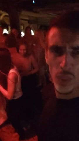 The video shows a man taking a selfie in what appears to be a crowded dance club. The lighting is dim with red hues. Other people are dancing and moving around in the background. The man's facial expressions change from surprise to a smirk. He is wearing a black shirt. The decor appears to be somewhat ornate.