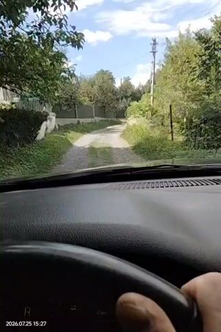 The video shows a first-person view from inside a car driving on a dirt road. The driver's hands are on the steering wheel. The dashboard shows the car is in drive (D) and has an odometer reading of 200439. Trees and a fence line the road, and the sky is partly cloudy. The timestamp on the video indicates July 25, 2026, at 15:27.