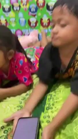 Two young children are lying down on a bed and looking at a mobile phone in their hands. The boy is wearing a black shirt and is on the right, while the girl wears a pink pajama top and is on the left. The bed is covered with a green patterned blanket, and behind them is a colorful patterned wall. The boy looks up at the camera, then back down at the phone. The girl smiles slightly as she looks at the phone.