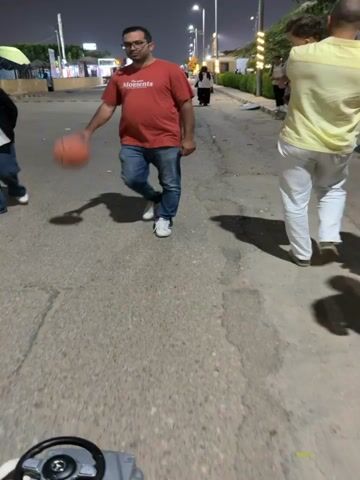 A man in a red t-shirt and blue jeans dribbles a basketball as he walks down a street at night. He is wearing glasses and white athletic shoes. In the background, other people are visible walking and standing along the side of the road. There are streetlights illuminating the scene. The perspective suggests the video is being filmed from a low-angle, possibly from a moving vehicle or a child's perspective.