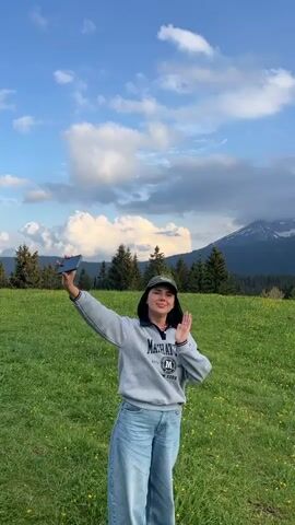A young woman stands in a flower-covered field, framed by trees and mountains, and takes a photo with her smartphone. The scene then shifts to a wide angle of the field, dotted with yellow flowers, extending towards small wooden houses nestled against a backdrop of imposing mountains partially shrouded in clouds. The sky is blue with scattered white clouds, and the view concludes with a glimpse of a white building with a red roof situated on the right.