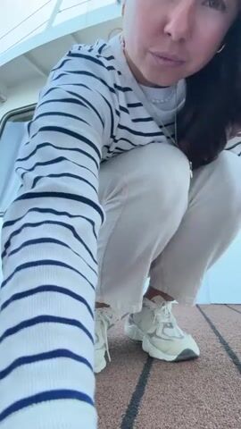 A woman with long dark hair, wearing a striped long-sleeved shirt and cream-colored pants, is crouching on a boat deck. She is looking down at her white sneakers. The deck is made of brown planks with dark lines. The woman then stands up and walks towards the side of the boat, leaning against the wall. She turns and smiles at the camera. The video ends with her in the same position.
