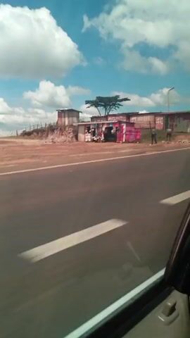 The video, taken from a moving vehicle, shows a roadside scene under a partly cloudy blue sky. The road is black asphalt with a white lane marker. The camera pans, revealing a series of small, makeshift buildings or stalls lining the side of the road. These structures appear to be constructed from corrugated metal and painted in various colors. People can be seen near these stalls. A large truck passes the camera at one point. The ground around the stalls is reddish-brown, contrasting with the green foliage of a tree in the background. Some parked vehicles are also visible near the stalls. A tall light pole stands by the roadside.