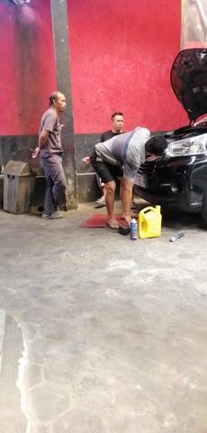 The video shows three men in what appears to be a garage or workshop. One man, wearing a grey and blue striped shirt and black shorts, is standing and working on the front of a black car with its hood open. Another man, wearing a dark t-shirt and light-colored shorts, is sitting on the floor next to the car, with his right hand near his mouth, possibly smoking. A third man, wearing a grey t-shirt and dark pants, is standing further away, looking towards the car. A yellow plastic container and a can are on the floor near the car. The environment is industrial, with red and black walls and a concrete floor. The video is recorded in landscape orientation but is presented in portrait, with the image rotated 90 degrees clockwise.