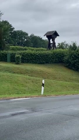 The video shows a bell tower on a grassy hill behind a hedgerow. A road is visible in the foreground, and the sky is overcast. The bell tower is made of dark wood and has a pitched roof. A bell is visible inside the tower. A white and black road marker is positioned on the left side of the road. The grass on the hill is green and appears to be well-maintained. Trees and bushes surround the bell tower, forming a natural backdrop. The overall impression is of a rural or semi-rural setting on a cloudy day.