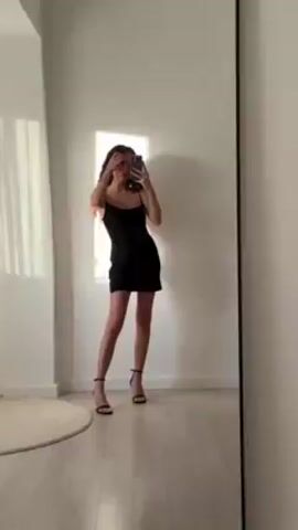 A woman with long dark hair stands in a room in front of a full-length mirror, taking a picture of herself with a phone. She is wearing a short black dress with spaghetti straps and black high-heeled sandals. She alternates between posing with her hand on her hip and brushing her hair behind her ear. The room is minimalist, with light-colored walls and wooden floors. A round rug is visible in the corner. A window allows sunlight to fall on the wall behind her.
