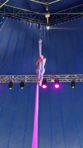 A person performs aerial silks inside a blue tent. The person, wearing a sparkly, light-colored costume, is suspended from a single point above by pink fabric. The person's body is contorted into a pose, with one leg extended and the other bent. The fabric hangs down in long folds. Stage lights, some green and some pink, are visible on a metal rig in the background.