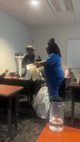 The video captures an indoor scene, likely a classroom or training room within a medical facility, featuring two women. Both women have dark skin and are dressed in scrubs. The woman on the left wears a black scrub top, glasses, and a patterned scrub cap, while the woman on the right is in blue scrubs with dark, curly hair.

They are standing next to a dark table, actively sorting through or examining the contents of several white bags placed on the table. The woman on the left is holding a bag open, and the woman on the right is reaching into another. One of the white bags appears to have red text, possibly "Christian Louboutin," while background audio mentions "Louis V's" and "Gucci," suggesting luxury items. A large, light-colored bag is on the floor next to the woman in blue.

In the background, a white medical scale with "DETECTO" printed on it is visible against a light-colored wall. The room contains several dark tables and chairs, indicative of a learning or meeting environment. The floor is covered with a patterned carpet. A clear water bottle with a black lid rests on a table in the foreground. The lighting suggests it is daytime. Casual conversation can be heard, including comments about the items in the bags.