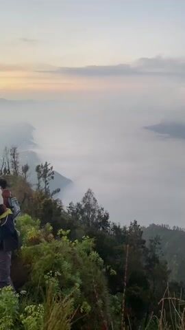 The video shows a landscape shrouded in thick fog, with only the peaks of mountains visible above the fog line. In the distance, a plume of white smoke rises from one of the mountain peaks. The foreground is filled with green vegetation, including trees and shrubs. The sky is a pale blue with some clouds. There are also some indistinct sounds of people talking in the background.