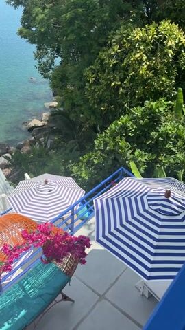 The camera pans up from a balcony with blue railings, showing a view of a seaside restaurant. The balcony has a lounge chair with a wicker headrest decorated with pink flowers. There are two large, blue and white striped umbrellas on the balcony. Below, the restaurant has white tables, blue chairs, and blue and white striped umbrellas. The restaurant is located on a rocky beach next to the ocean, which is clear and blue-green. Trees surround the restaurant, and a tall tree is visible to the left of the frame.