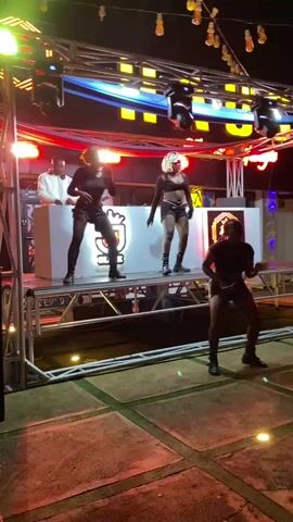 Three women are dancing on a raised stage in front of a DJ booth. The woman on the far left is wearing a white shirt and black pants. The woman in the center is wearing a black crop top and black shorts. The woman on the far right is wearing a black dress. The DJ is standing behind the booth. The stage is lit with colorful lights. The background appears to be an outdoor venue with string lights overhead.