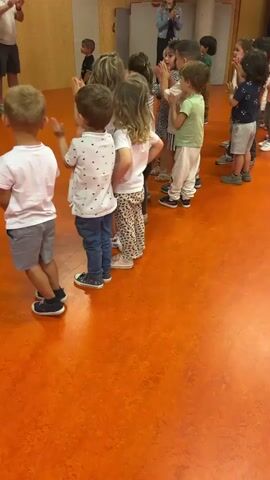 A group of young children are standing in a line on an orange floor and clapping their hands. The children are wearing casual clothing. The environment appears to be a classroom or indoor play area. The focus is on the children clapping.