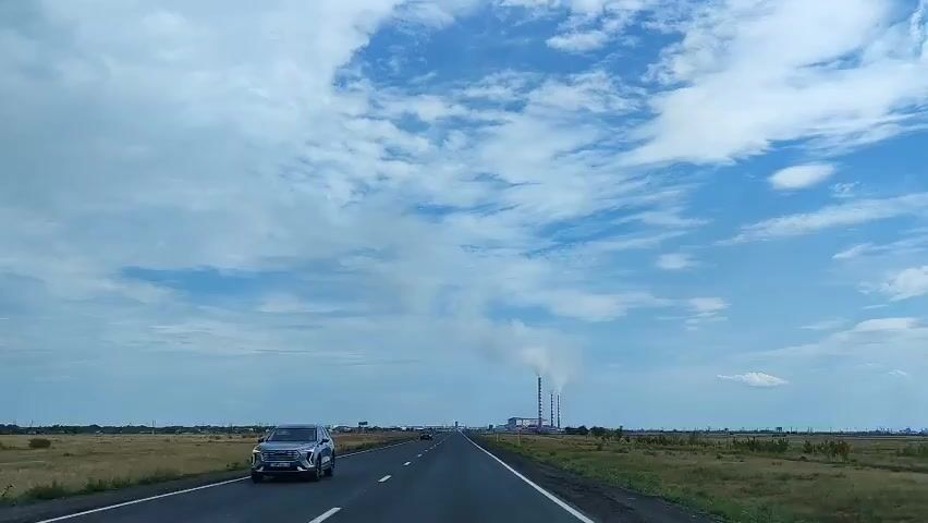 The video shows a highway stretching into the distance under a partly cloudy blue sky. On the right side of the road, a sign reads "АКСУ". In the distance, a large industrial complex with several tall smokestacks is visible, emitting plumes of white smoke. The landscape on either side of the highway is flat and covered in dry grass. A few cars are seen on the road, and one car is passing the camera from the opposite direction. The overall impression is a journey through an open, rural landscape towards an industrial area.