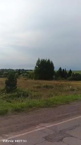 This is a video of a landscape shot from a moving vehicle. The video begins with a view of a rural road with green grass and trees on either side. The sun is visible in the sky, casting a bright light on the scene. As the video progresses, the vehicle continues to move, and the landscape changes slightly. The trees become more prominent, and there are some rolling hills in the distance. The sky is partly cloudy, with patches of blue visible. The video ends with a final shot of the landscape, showing a continued view of the rural scenery. The watermark "Infinix HOT 50" is visible in the bottom left corner of the screen throughout the video.