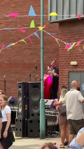 A medium shot captures a street performance outside a brick building. A person dressed in a black outfit with a bright pink skirt and a red headdress stands on a raised platform, playing a double bass. Two large speakers are stacked to the left of the platform. A microphone is positioned in front of the musician. Colorful triangular flags are strung across the scene, attached to a teal pole that runs vertically in the foreground. Several people are visible in the foreground, facing the performance. On the right, a man with a beard and a white shirt stands partially turned away from the camera, while a woman with blonde hair in a striped top stands next to him. On the left, a young girl with blonde hair in a white top and black skirt is partially visible. The background is a brick wall with a doorway. The ground appears to be pavement.