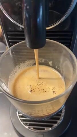 A close-up video captures a coffee machine actively brewing coffee into a clear glass mug. A steady, light brown stream of coffee, topped with a thick layer of crema and small bubbles, descends from a black dispenser spout into the mug. The mug rests on a metallic grid, part of the coffee machine's drip tray. The machine itself is dark-colored, with a textured surface visible in the blurred background. The scene suggests an indoor setting, focusing solely on the coffee preparation process. No people or visible text are present. This activity is taking place, by context, in Chambœuf, France.