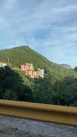 The video shows a view from a moving vehicle looking towards a hillside town. There are several buildings, mostly multi-story structures with red or white roofs, nestled amongst lush green trees and vegetation. The buildings appear to be residential or possibly hotels, with balconies visible on some of them. The sky is blue with some light cloud cover. A yellow guardrail is in the foreground, indicating the perspective is from a road. The overall impression is a scenic view of a coastal or mountainous settlement.