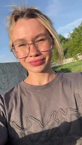 A blonde woman with glasses smiles at the camera in an outdoor setting. Her hair is messy and she wears a gray T-shirt. Behind her, other people are visible in the background, some appearing to be working in a garden or yard. There are also a few structures, like sheds or fences, in the background, suggesting a rural or suburban setting. The weather appears sunny, with a clear blue sky.