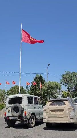 The video shows a bright, clear, blue sky. A large red flag with a yellow symbol in the center flies from a tall white flagpole. Several smaller red flags are visible in the background. In the foreground, two vehicles are parked side-by-side. The vehicle on the left is a light-colored SUV with a spare tire mounted on the back. The vehicle on the right is a darker-colored SUV, appearing dusty. Trees and some electrical wires are also visible in the background.