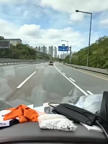 THE VIDEO SHOWS A FORWARD-FACING VIEW FROM A VEHICLE TRAVELING ON A MULTI-LANE HIGHWAY. IN THE FOREGROUND, ON THE DASHBOARD, ARE SEVERAL ITEMS INCLUDING ORANGE GLOVES, A BLACK FACE MASK, AND A PLASTIC CONTAINER WITH WHAT APPEAR TO BE UMBRELLA COMPONENTS OR SIMILAR LONG, THIN OBJECTS. IN THE MIDGROUND, OTHER VEHICLES ARE VISIBLE ON THE HIGHWAY, INCLUDING A SMALL TRUCK CARRYING LONG, CYLINDRICAL OBJECTS ON ITS ROOF. A SPEED LIMIT SIGN INDICATING 70 IS VISIBLE. IN THE BACKGROUND, NUMEROUS HIGH-RISE APARTMENT BUILDINGS ARE SITUATED ON A HILLSIDE, AND TREES LINE THE SIDES OF THE HIGHWAY. THE SKY IS OVERCAST WITH CLOUDS. THE ROAD APPEARS TO BE WET, REFLECTING THE SKY AND SURROUNDINGS. THERE IS A LANE MARKER INDICATING A RIGHT TURN OR EXIT.