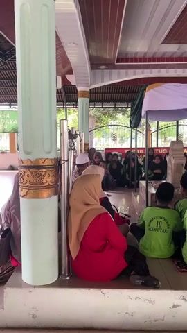 A group of people, mostly children wearing green shirts and black caps, are seated on the floor facing a tomb. The tomb is covered by a white and green canopy. Some adults are also present, one in a red shirt and hijab, and another man wearing a white cap and shirt sitting near the tomb. The scene appears to be outdoors, possibly at a religious site, with a tiled ceiling and large pillars visible. The children seem to be listening to the man near the tomb.