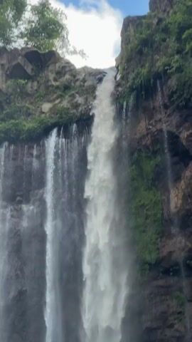 The video shows a tall waterfall cascading down a rocky cliff face. The water is white and foamy as it falls, creating a misty spray. The cliff is covered in green vegetation in some areas, and bare rock in others. The sky is visible at the top of the frame, with some clouds. The overall impression is one of natural beauty and power.