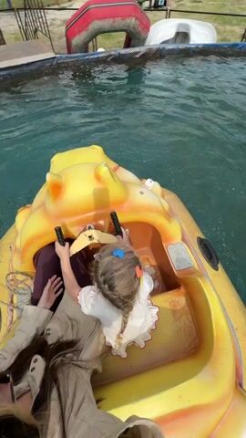 Here's a description of the video:

The video shows a woman and a young child, seemingly a girl, riding in a small, yellow, hippo-shaped paddle boat on a pool of water. The child, wearing a white dress and a blue hair clip, is holding onto the boat's steering wheel. The woman is wearing sunglasses and a light-colored jacket. The water in the pool is a light blue color. In the background, there are other amusement park attractions and people walking around. The camera is positioned from the woman's perspective in the boat, looking down at the child and the water. The boat moves gently on the water.