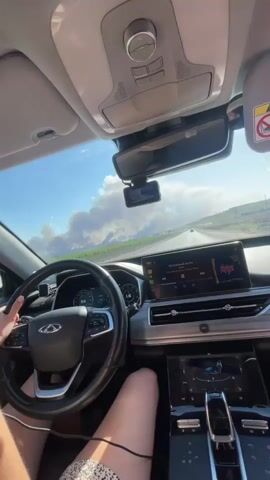 The video begins with a first-person perspective from inside a car. The camera is mounted facing forward and slightly upwards, capturing the dashboard, steering wheel, and the view through the windshield. A person's hands are visible on the steering wheel, indicating they are driving. The dashboard features a digital speedometer and a large infotainment screen displaying what appears to be music playback. The windshield shows a highway stretching into the distance under a bright, partly cloudy sky. In the far distance, a large plume of smoke rises from what appears to be a fire. The interior of the car is well-lit, and the overall impression is of a journey on a sunny day with a distant environmental concern. The video is very short, with the timestamp indicating it is only a few seconds long.