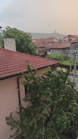 The video shows an aerial view of a residential area with houses and trees. The houses have red tiled roofs and some have chimneys. The trees are green and leafy, and appear to be swaying in the wind. In the background, there is a hill with a tall structure on top. The sky is hazy and overcast, suggesting it might be raining or there could be dust in the air. There are no people visible in the video, and the overall scene depicts a somewhat unsettled weather condition in a quiet neighborhood.