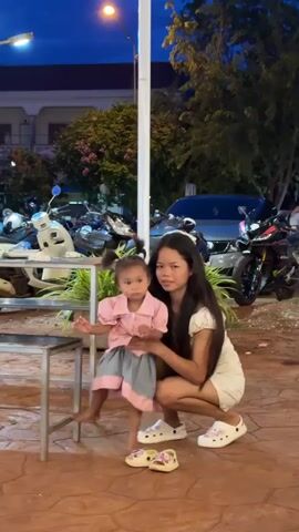 A young girl with dark hair in pigtails, wearing a pink button-down shirt and a grey skirt, stands near a metal table. A young woman with long dark hair, wearing a white dress, kneels beside her, holding the girl's left foot. Both are wearing white Crocs with pink floral patterns. The woman appears to be putting on or adjusting the girl's shoe. The scene is outdoors at dusk, with trees, buildings, and parked motorcycles in the background.