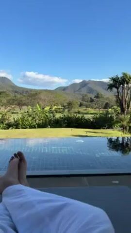 The video shows a person lying on a lounge chair next to a pool. The person is wearing white pants and their bare feet are visible. The pool is an infinity pool that overlooks a green field and mountains in the background. The sky is blue with a few clouds. There are trees and other plants in the foreground.