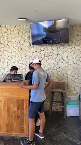 The video shows a hotel reception area.  Two young men stand at the wooden reception desk, talking to a female receptionist. The receptionist looks at a tablet on the counter. Behind the desk is a wall covered with light gray stones. Above the desk, two flat-screen televisions display images of the hotel and its surroundings. A wooden stool is visible near the desk, along with informational pamphlets. The floor appears to be made of dark-colored tiles.