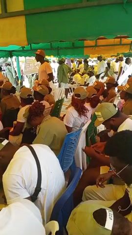The media file depicts a gathering of young adults, likely participants in a National Youth Service Corps (NYSC) program, as indicated by the "NYSC" text visible on a white t-shirt worn by a man standing on the right. The scene is set outdoors, under a green and yellow tent, suggesting it is a daytime event. The participants are predominantly dressed in light green uniforms, including shirts and trousers, and many are wearing matching green caps.

The individuals are seated on plastic chairs, some in rows, and appear to be engaged in various activities. Some are looking at their phones, while others are reading documents or interacting with those around them. The overall atmosphere seems relaxed and informal. The presence of multiple people, the tent structure, and the uniform attire strongly suggest an organized event or gathering related to the NYSC program.

In the foreground, several individuals are captured in detail, including a man wearing glasses and earphones, and another with a white shirt and a black strap. The background shows a larger crowd of people, further emphasizing the scale of the event. The lighting suggests it is daytime, possibly midday or afternoon, with no specific indicators of the weather conditions, though the tent suggests a need for shade. There are no overt signs of distress or urgency; the people appear to be waiting or participating passively in the event. The setting seems to be a community area or a designated event ground in Ajiwa, Nigeria.