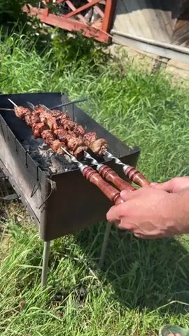 A person is cooking shish kebab on a charcoal grill in a grassy yard. Three skewers loaded with chunks of meat are seen on top of a simple metal grill. The person is holding the skewers with their hand. Smoke rises from the coals below, and the meat is sizzling. The ground around the grill is covered in green grass, and a wooden structure is visible in the background. The skewers have twisted metal designs and wooden handles.