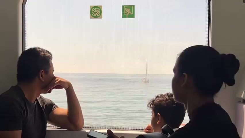 The video shows a family on a train ride, looking out the window at the ocean. A man is on the left, his arm resting on the windowsill and his hand supporting his chin. A woman is on the right, and between her and the man is a young boy. They are all looking out the window, which shows a bright, sunny day over the water. There are two signs on the window. In the foreground, a hand holds a smartphone. The train appears to be moving, as indicated by the slight blur of the scenery outside the window.