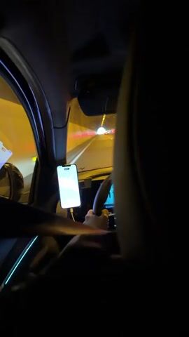 The video shows an interior view of a car driving through a tunnel at night. The driver is visible holding the steering wheel, with a smartphone mounted to the left of the wheel displaying a navigation app. The speedometer on the dashboard shows the car's speed fluctuating. The tunnel is brightly lit with yellow lights, and other cars can be seen in the distance. The viewpoint appears to be from the backseat.