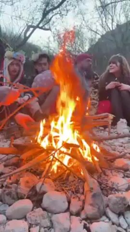A group of five people sits around a campfire in a rocky outdoor setting. The fire is the focal point, burning brightly in the center of the frame. The people are positioned around the fire, seemingly engaged in conversation or simply enjoying the warmth. They are dressed in casual clothing, some with headwear that suggests a festive or nature-themed event. The environment consists of a rocky ground with trees and vegetation in the background, indicating a natural setting like a forest or riverside. The sky is overcast, creating a soft, diffused light. One person stands up from the group and walks away from the fire.