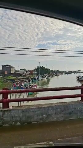 The video shows a view from a vehicle, looking out over a body of water with numerous boats. The water appears to be a river or canal, with a concrete structure and red railing in the foreground. Beyond the railing, several small fishing boats are docked or anchored. Some of the boats are colorful, with red, green, and white exteriors. In the background, there are trees and buildings, suggesting a coastal or riverside village. The sky is partially cloudy, with a bright, hazy sun. The camera appears to be moving, indicated by the slight shift in perspective as the video progresses. There are no people clearly visible in the video.