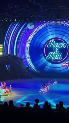 The video captures a live show scene with performers on an ice-covered stage. The backdrop is dominated by a large LED screen displaying a graphic of a record player and the words "Rock & Roll." The stage lighting is blue, creating a cool ambiance. Several performers are seen riding unicycles with illuminated wheels, while another performer stands nearby in a red outfit. In the background, a car decorated with flowers is visible. A group of spectators is seen in the foreground, silhouetted against the stage lights. The camera angle is static, focusing on the stage action.