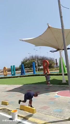 The video pans across a public space in La Guaira, Venezuela, on a bright, sunny day. The most prominent subjects are large, colorful, three-dimensional letters spelling out "LA GUAIRA." The letters are red, orange, teal, blue, and red, arranged in a row. Behind the letters, a grassy area is visible, with a statue of a person on horseback situated in the middle. Further in the background, trees and palm trees dot the landscape, with the ocean visible in the far distance. The sky is clear and blue, suggesting pleasant weather.

In the foreground, a paved walkway with a colorful, mosaic-like pattern leads the viewer's eye towards the lettering. Yellow cylindrical bollards are placed along the edge of the walkway, one of which is partially visible at the bottom left of the frame. The overall impression is of a vibrant tourist attraction or public plaza designed to welcome visitors and showcase the city's name. The clear weather and bright colors evoke a feeling of cheerfulness and openness. No people are clearly visible in the frame. The visible text at the bottom of the screen reads "Infinix HOT 50 Pro."