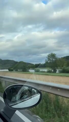 The video shows a view from inside a car, looking out the side window. The landscape appears to be rural, with rolling hills covered in trees and vineyards. A body of water, possibly a river or lake, is visible in the middle ground, with fields of harvested crops in the foreground. There are several road signs visible, including speed limit signs and directional signs. The sky is overcast with dramatic clouds. The interior of the car is partially visible, including a side mirror reflecting the landscape. The video appears to have been filmed while the car was in motion.
