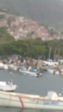 The video shows a harbor with several boats docked. In the background, a hillside is covered with densely packed houses. There are trees and some structures on the shore, along with a white truck. People are visible near the boats and on the shore. The water appears calm. Some of the boats have outboard motors. One boat in the foreground has the name "LAS CATALINAS" on its side. The sky is overcast and the overall scene suggests a coastal settlement in a hilly region.