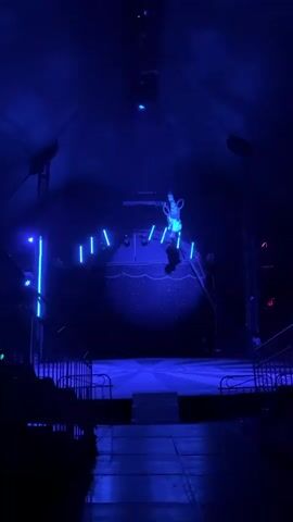 The video shows a person performing a hanging acrobatic act at a circus. The act is illuminated by blue and yellow lights within a dark tent. The person is hanging upside down, with their body contorted in a flexible pose. The circus set includes a stage with a patterned floor and a large, round prop decorated with stars. The stage is surrounded by metal railings and stairs.  A dramatic, possibly romantic, song plays in the background.