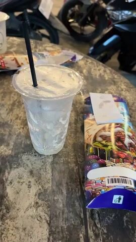 This video captures a static shot of a table in what appears to be an outdoor or semi-outdoor setting in Gampongjawa, Indonesia. The main subjects are a cold beverage and a food package placed on a light grey, textured wooden table.

In the foreground, a clear plastic cup, visibly covered in condensation, contains a white or light-colored iced drink with a black straw. To its right, a colorful rectangular food package, predominantly blue and purple, features an illustration of a wrap or kebab on its front. The package also has a visible barcode and a trash/recycling symbol. On the left side of the frame, another clear plastic cup with a partially visible logo that reads "FORETTE" is also on the table.

The background is softly blurred, showing several parked motorcycles, suggesting the location is near a street or parking area. The lighting indicates it is daytime. There is no direct human activity shown in the video; however, ambient background sounds are audible, suggesting general activity or traffic in the vicinity. The condensation on the cup implies a warm ambient temperature.