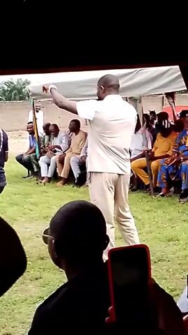 A man in a light-colored shirt and pants gestures with his right hand while facing a seated audience. He appears to be speaking. Several people are seated on a bench behind him, and others are standing. A camera on a tripod is positioned to the left of the scene, and a speaker is on the ground next to it. The setting appears to be outdoors, with grass and a fence visible in the background. The video is shot from a first-person perspective, with a hand holding a red-cased smartphone visible in the foreground.
