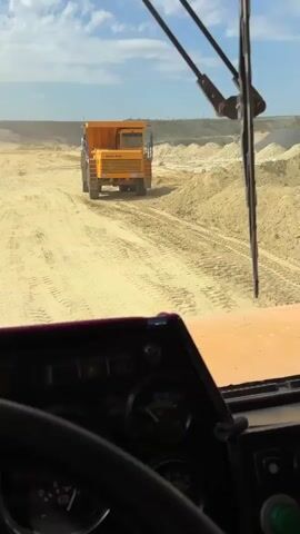 The video is filmed from the perspective of the driver inside a vehicle, looking out through the windshield. A large, orange dump truck is visible in the distance on a dirt road. The dump truck is moving away from the camera. The surrounding environment appears to be a quarry or construction site, with piles of sand or dirt visible on either side of the road. The sky is cloudy. The interior of the vehicle shows a dashboard with gauges and buttons. A windshield wiper is present in the foreground.