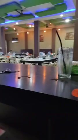 The video shows an interior view of a restaurant or dining establishment. The focus is on a dark-colored table in the foreground with a set of keys and some liquid spills. To the right of the keys is a glass with a black straw and some green liquid, possibly a beverage. In the background, multiple tables are set with white tablecloths, water bottles, and glasses. The ceiling features green and blue lighting, and there are ceiling fans visible. The overall impression is of a quiet or empty dining area, possibly during off-hours or before a rush. The lighting is somewhat dim, with reflections visible on the table surface.