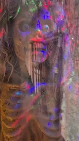 The video shows a zombie prop behind a glass barrier, illuminated with multicolored lights. The zombie has long, curly hair, a skeletal chest, and a visible set of teeth. The lighting creates a blurred, ethereal effect on the prop.