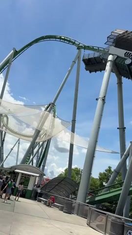 The video shows a green roller coaster at what appears to be a theme park, shot from a ground-level perspective. The roller coaster's track is supported by grey beams. The video captures the moment a full roller coaster cart ascends a section of the track that is constructed to look like a giant cannon. In the background, people are seen walking and sitting near a small building. The sky is bright blue with some clouds. A recycle bin can be seen in the foreground.